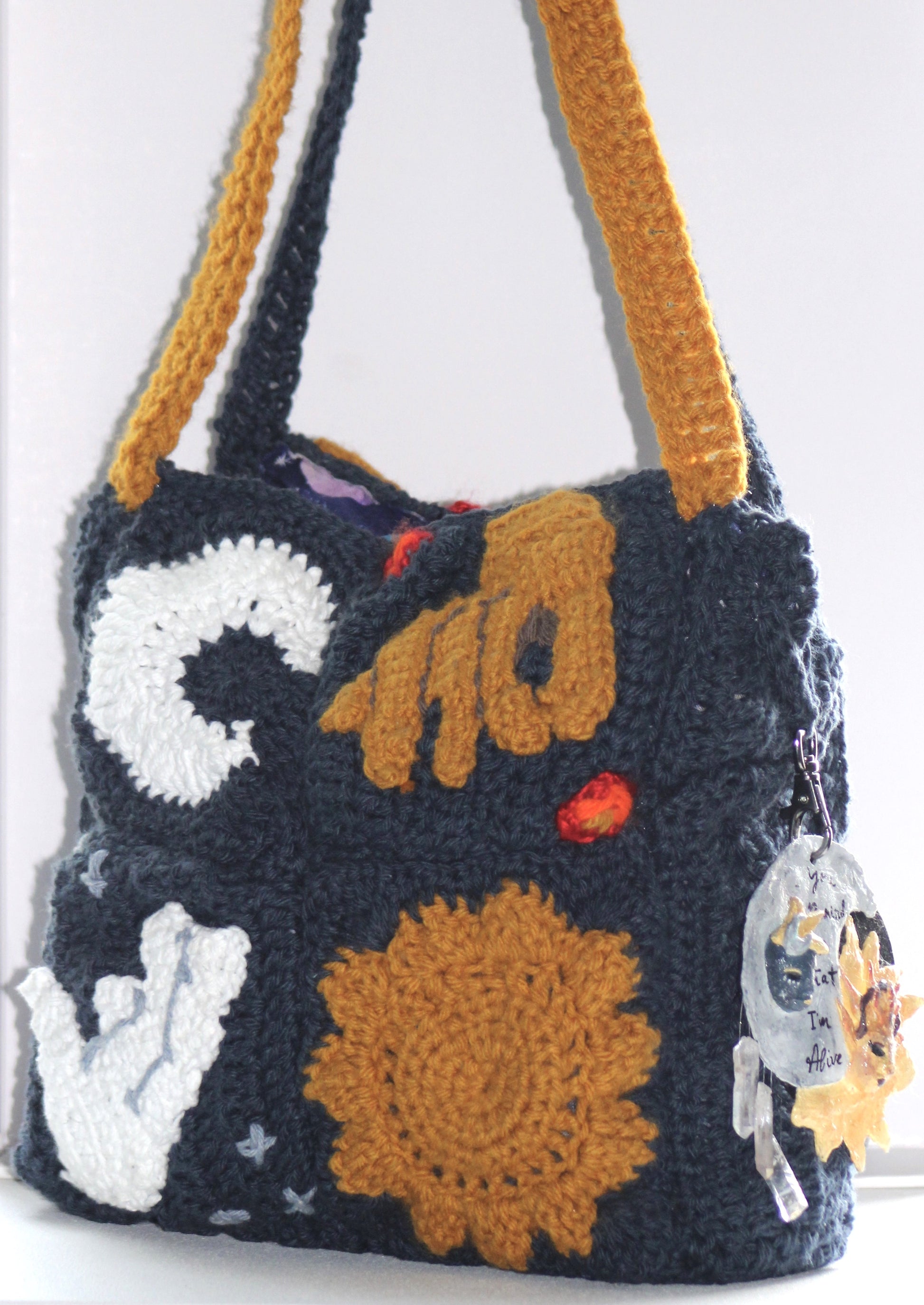 Front view of navy blue crochet purse made up of four granny squares. The top left square has a white crescent moon, the top right has a golden hand reaching toward the bottom left, the bottom left has a white hand reaching toward the top right, and the bottom right has a golden sun. There are also three stars in the two corners above and below the white hand granny square and one flame in the two corners above and below the golden hand granny square. This handbag has two straps, one blue and one golden.