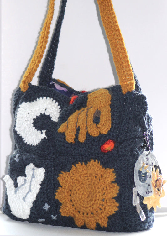 Front view of navy blue crochet purse made up of four granny squares. The top left square has a white crescent moon, the top right has a golden hand reaching toward the bottom left, the bottom left has a white hand reaching toward the top right, and the bottom right has a golden sun. There are also three stars in the two corners above and below the white hand granny square and one flame in the two corners above and below the golden hand granny square. This handbag has two straps, one blue and one golden.