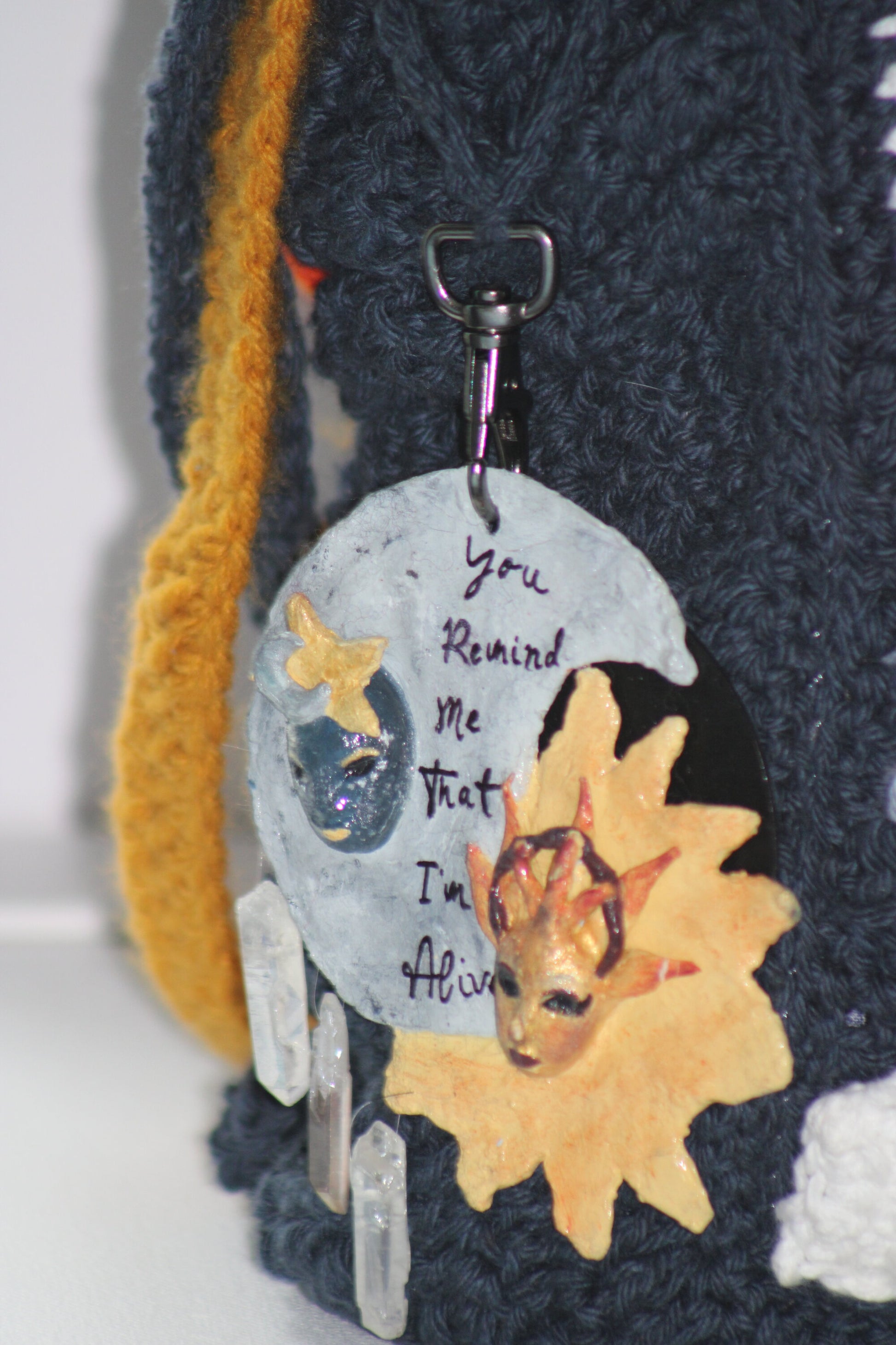 This handbag also has an attachable keychain on the side, which contains a blue male moon and star masquerade mask and a yellow female sun masquerade mask. There are also three crystals hanging to the bottom left and the quote "You Remind Me That I'm Alive" written between the two masks. 